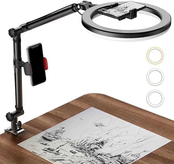 A black LUOLED 10″ Ring Light with a phone holder clamped to a wooden desk, illuminating a drawing of a cityscape. A smartphone is mounted in the ring light’s holder.