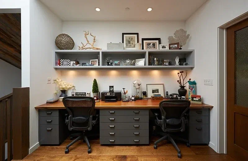 Home office with layered lighting: overhead spotlights, shelf lighting, and desk lamps. Two ergonomic chairs and a long shared desk.